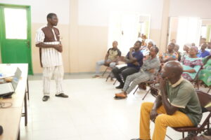 Man in traditional attire speaking to an attentive audience in a bright room with a green door on the left.