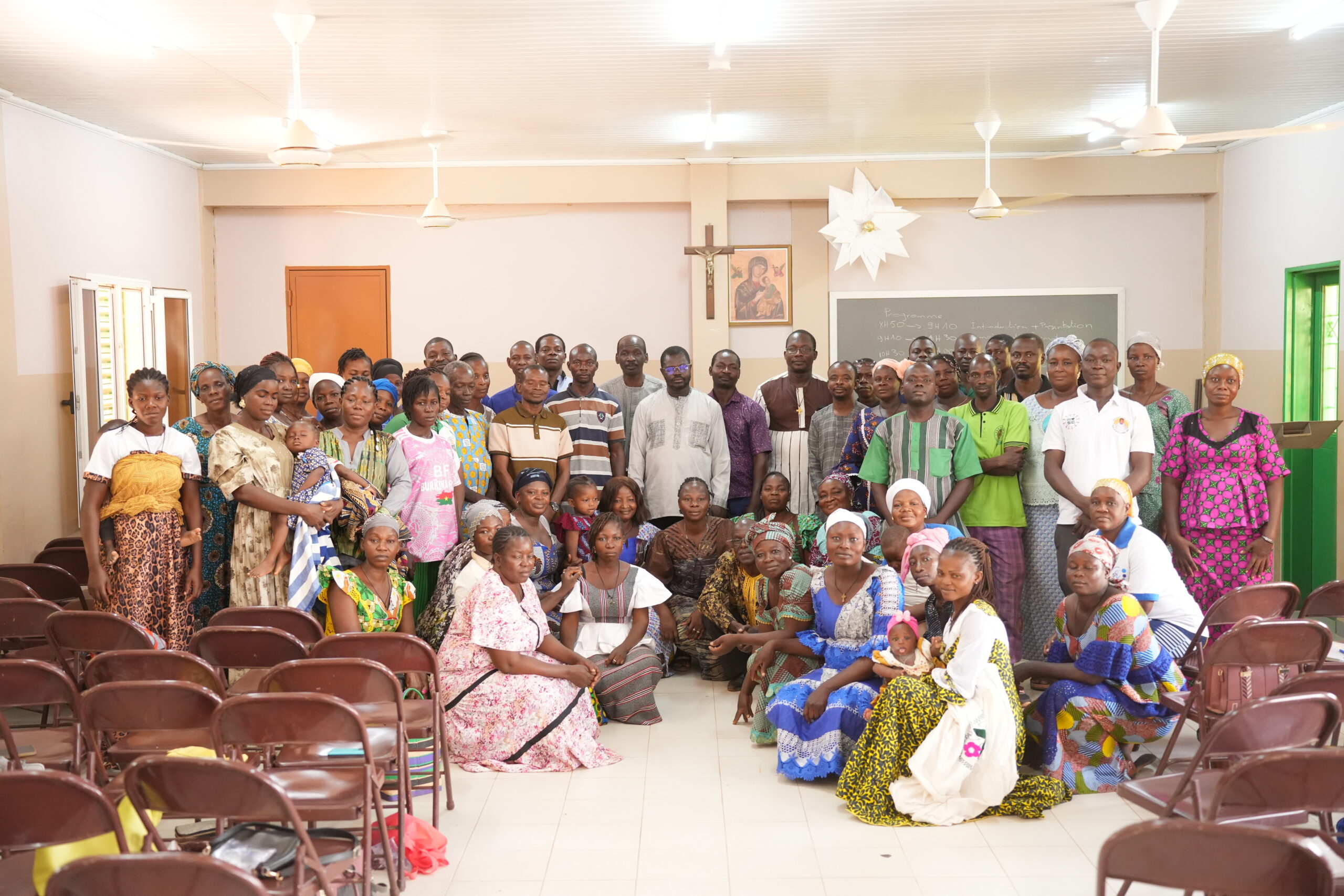 Large diverse group of people in a church hall posing for a group photo, many wearing bright traditional clothing; crucifix and religious icon on the wall behind them.