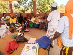 Group of women with infants sitting in a semicircle under a shaded area for a community health workshop.