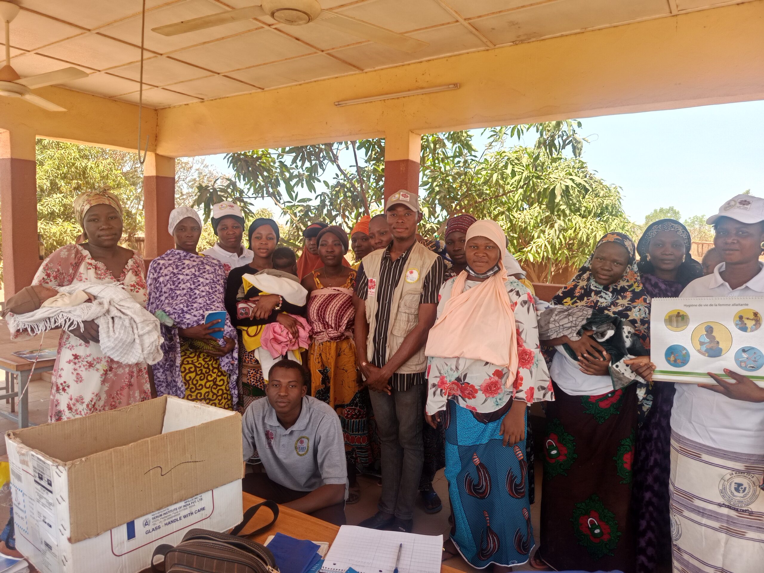 Group of women and children under a sheltered area during a community health outreach, volunteers handing out blankets and baby items.