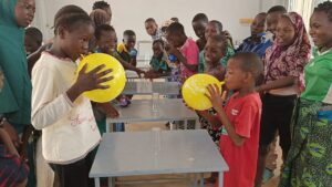 Group of children in a classroom crowd around metal desks, two kids blowing yellow balloons as others watch with smiles.