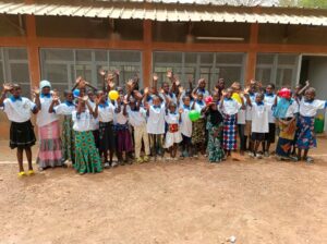 Group of smiling children in white shirts waves hello outdoors in front of a classroom building; some hold colorful balloons.