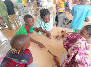 Group of children and an adult facilitator gather around a table, pointing to rectangular boxes drawn on a large sheet of brown paper for a classroom activity.
