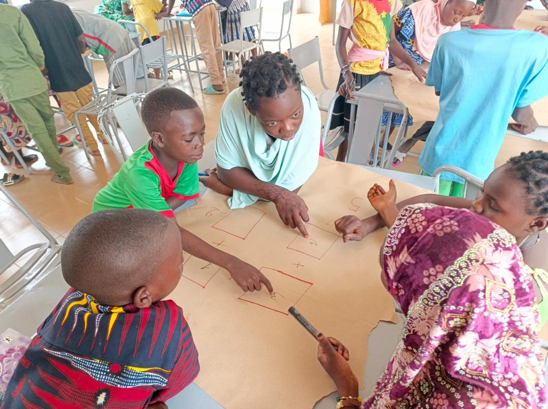 Group of children and an adult facilitator gather around a table, pointing to rectangular boxes drawn on a large sheet of brown paper for a classroom activity.