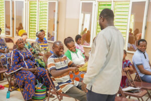 Community group seated in a waiting room or hall, smiling as a man stands and speaks to them.