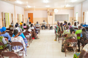 Crowded room of people in colorful attire seated for a presentation, with a speaker at a table and a chalkboard at the front.