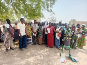 A group of villagers, mostly women and children, gathered under a tree as a woman in a red dress speaks to them, holding a paper in her hand.