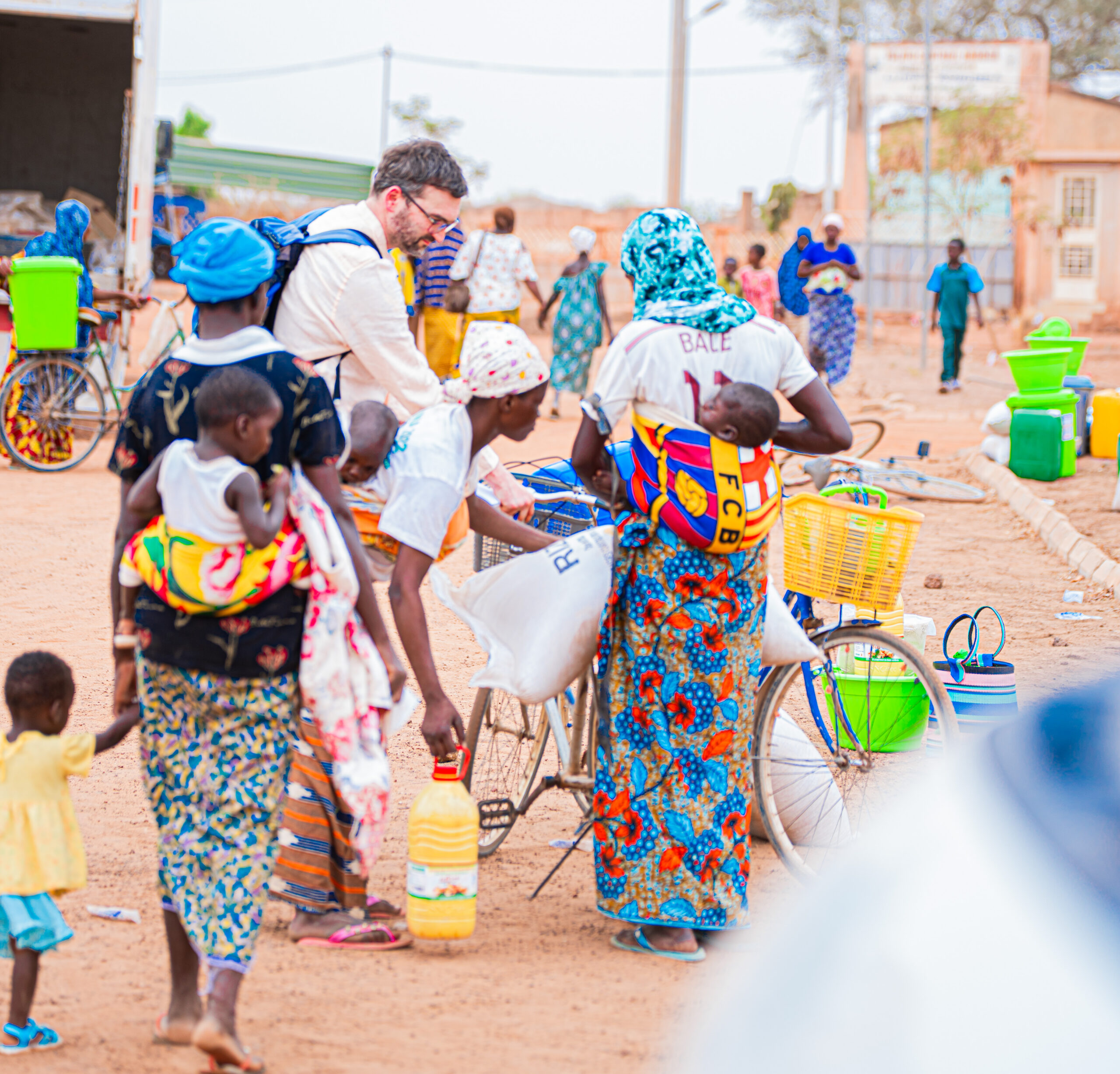 Group of people in colorful clothing gathered on a dirt street with bicycles and yellow water jugs nearby in a village setting.