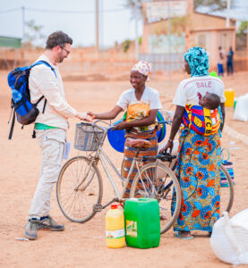 Man shakes hands with a smiling girl beside a bicycle in a sunny, dusty outdoor area; others in colorful clothing nearby with water containers nearby.