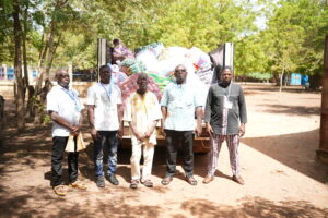 DSC04679 - Tous pour l'homme, chacun pour le faible Five men stand in front of a truck loaded with colorful bags outdoors, sunlit with green trees in the background.