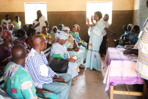 DSC04697 - Tous pour l'homme, chacun pour le faible A woman in a blue patterned dress speaks to a seated audience in a community hall as people listen attentively.