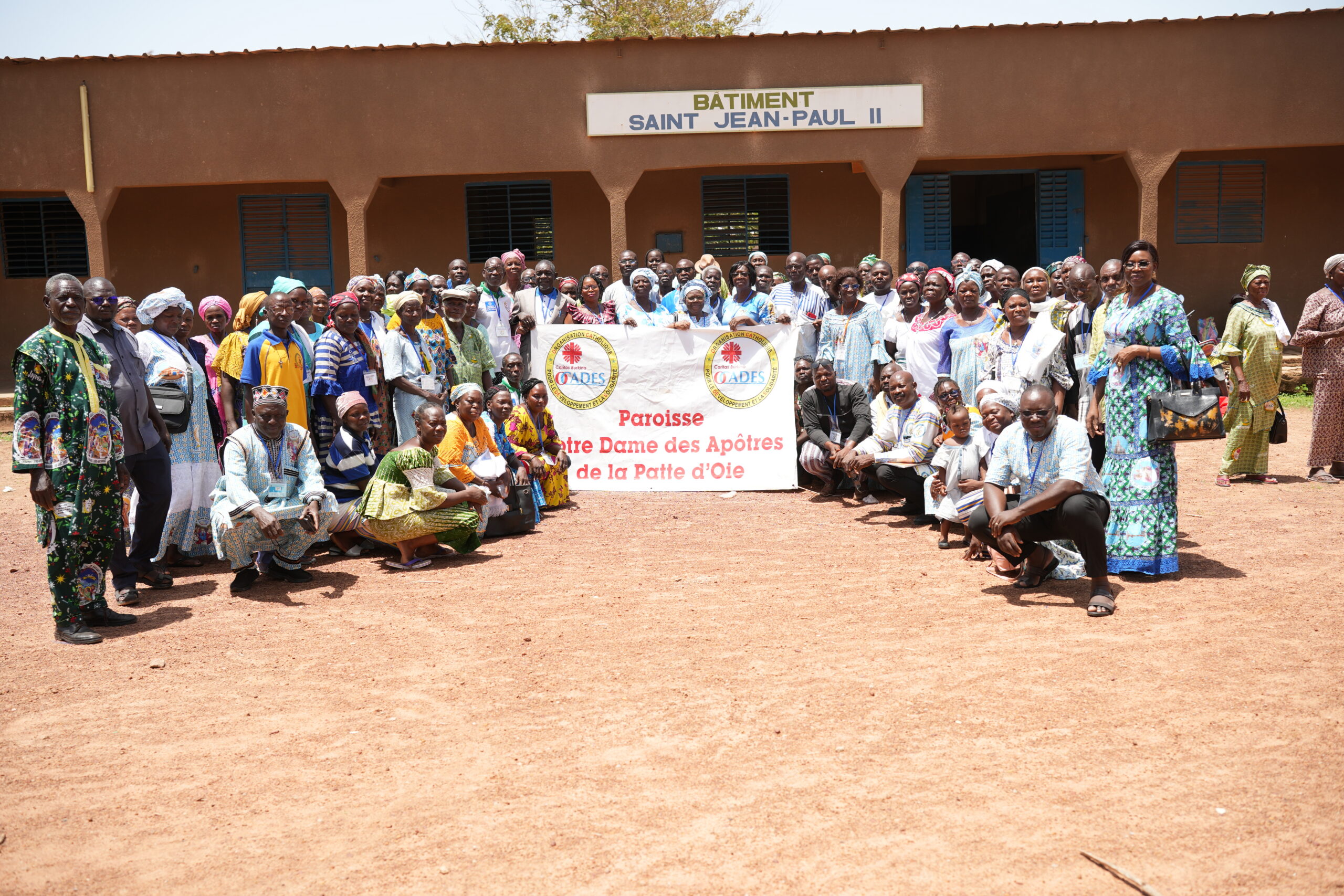 Large group of people posed outdoors in front of a brown building labeled Saint Jean-Paul II, holding a parish banner.