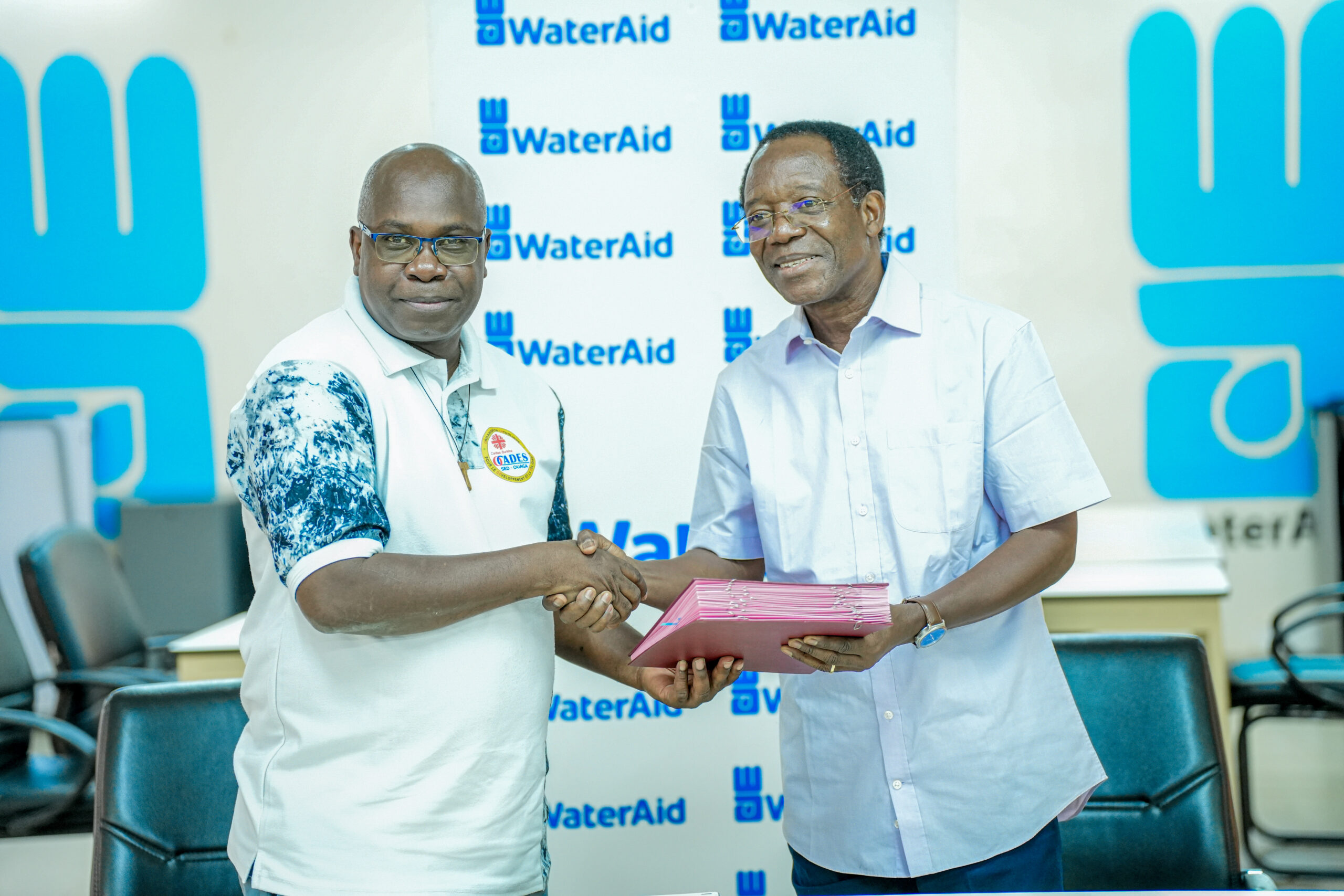 Two men shaking hands in front of a WaterAid backdrop; one holds a pink folder in their other hand.