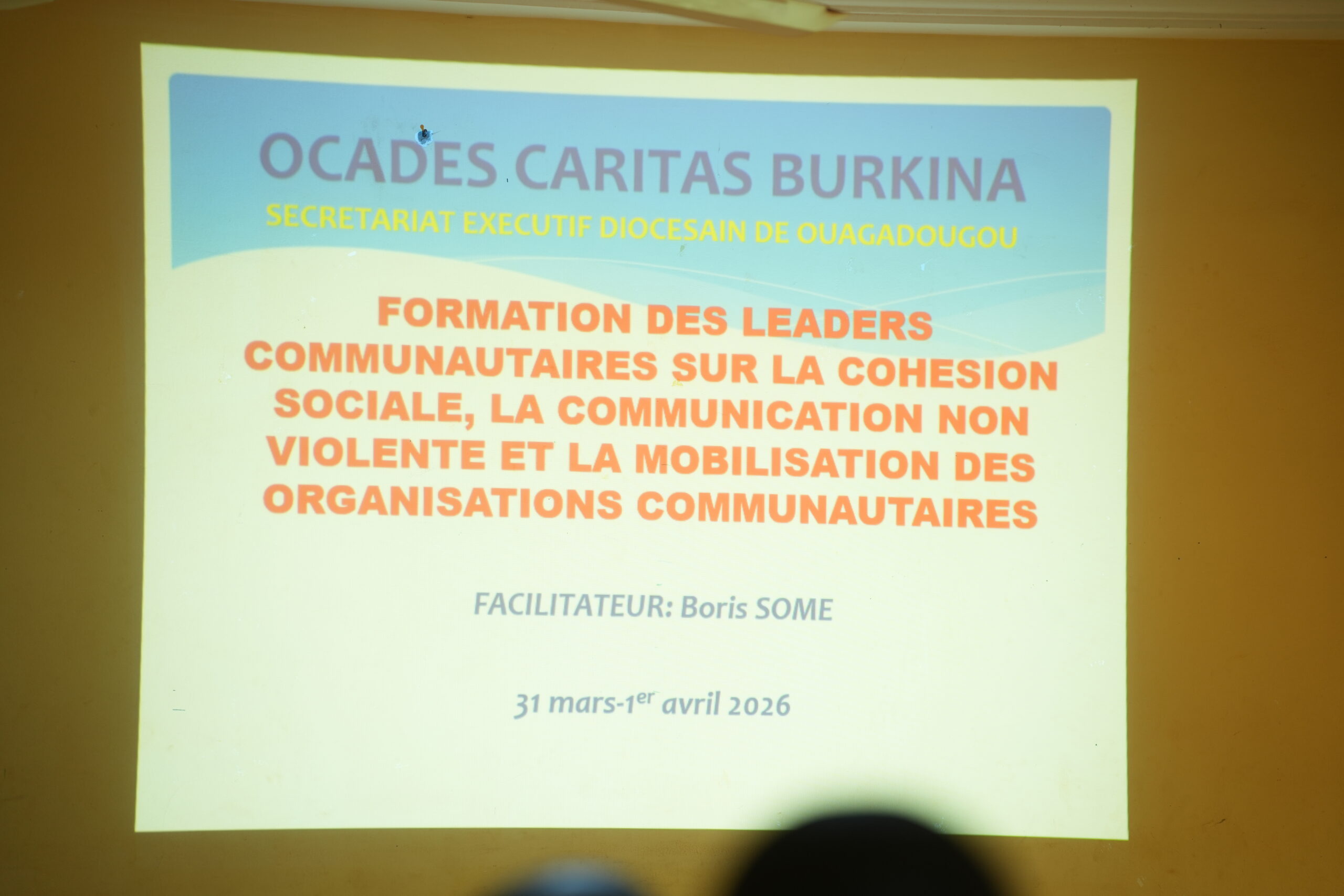 Projection of a slide reading 'OCADES CARITAS BURKINA' in blue, with yellow subtitle and orange bold title about leadership training on social cohesion; bottom shows 'Facilitateur: Boris SOME' and dates.