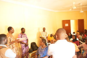 Group of people in colorful traditional clothing gathered indoors for a meeting or event in a bright yellow-lit room.