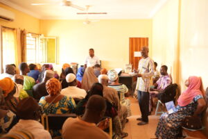 A crowded classroom-like hall with a speaker at the front addressing a diverse audience in colorful traditional clothing.