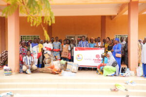 Group of men, women, and children posing for a charity donation event outside a peach-colored building, with bags, boxes, and supplies in the foreground and a banner displaying organizational logos behind them.