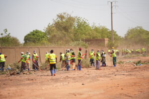 Group of people in bright safety vests sweeping and picking up litter in a dusty outdoor area near a wall, wearing masks and boots.