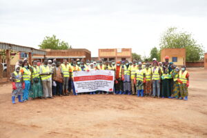 Large group of people in yellow safety vests posing with a banner at a dusty construction site, outdoors.