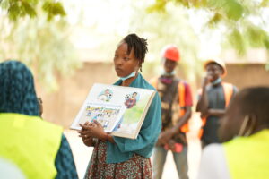 Woman in teal clothes with mask under chin presents a colorful illustrated workbook to a group of workers in high‑visibility vests outdoors.