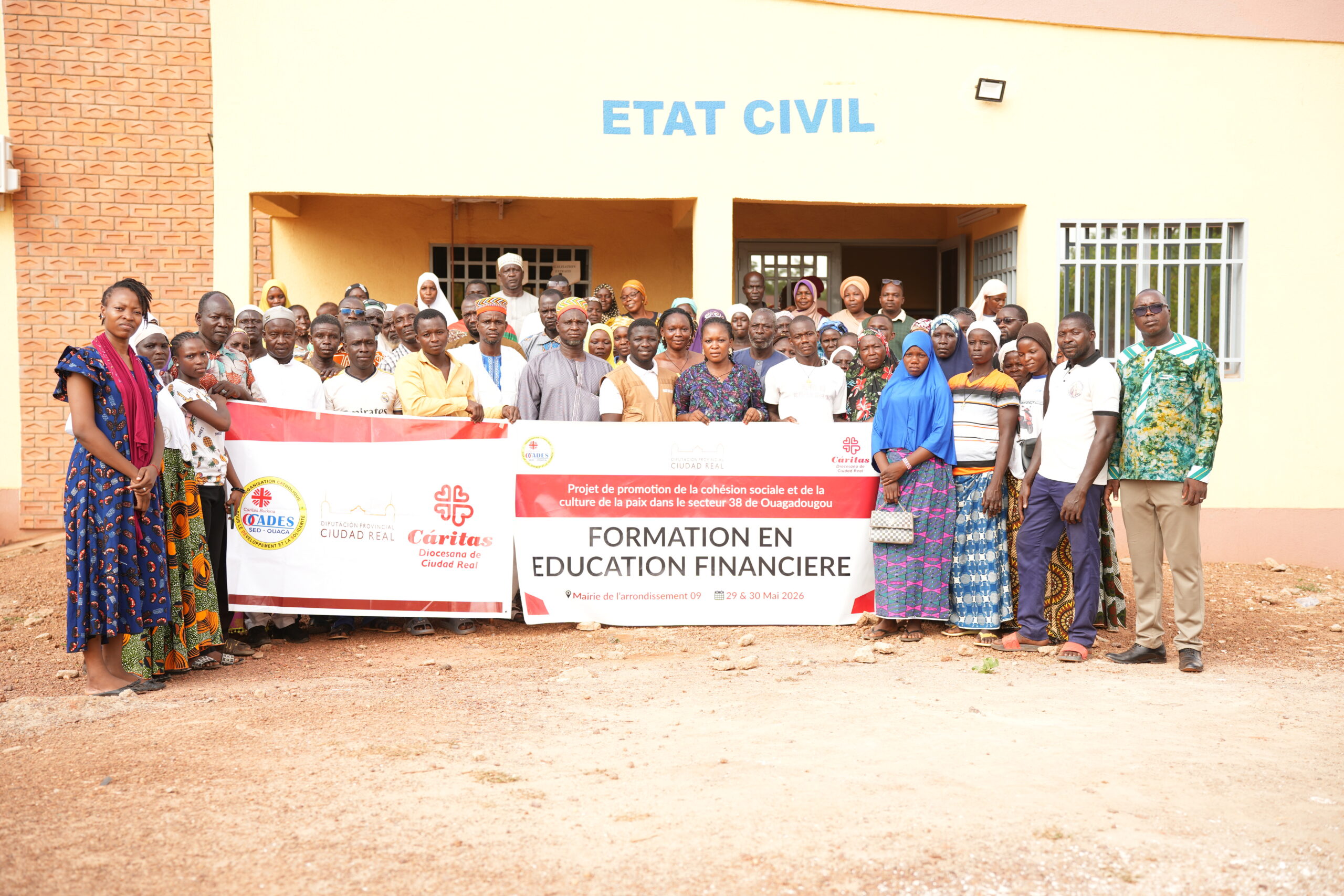 Group of men and women posing in front of a yellow building with 'ETAT CIVIL' sign, holding a banner for a social cohesion and financial education program.