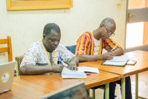 WhatsApp Image 2026-07-17 at 193700 1 - Tous pour l'homme, chacun pour le faible Two men sit at a wooden table studying documents; one wears a patterned shirt and the other a red‑yellow striped shirt, both writing notes.