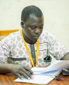 WhatsApp Image 2026-07-17 at 193701 1 - Tous pour l'homme, chacun pour le faible Man focused on writing in a notebook at a table, wearing a patterned shirt and an orange lanyard with a badge visible.