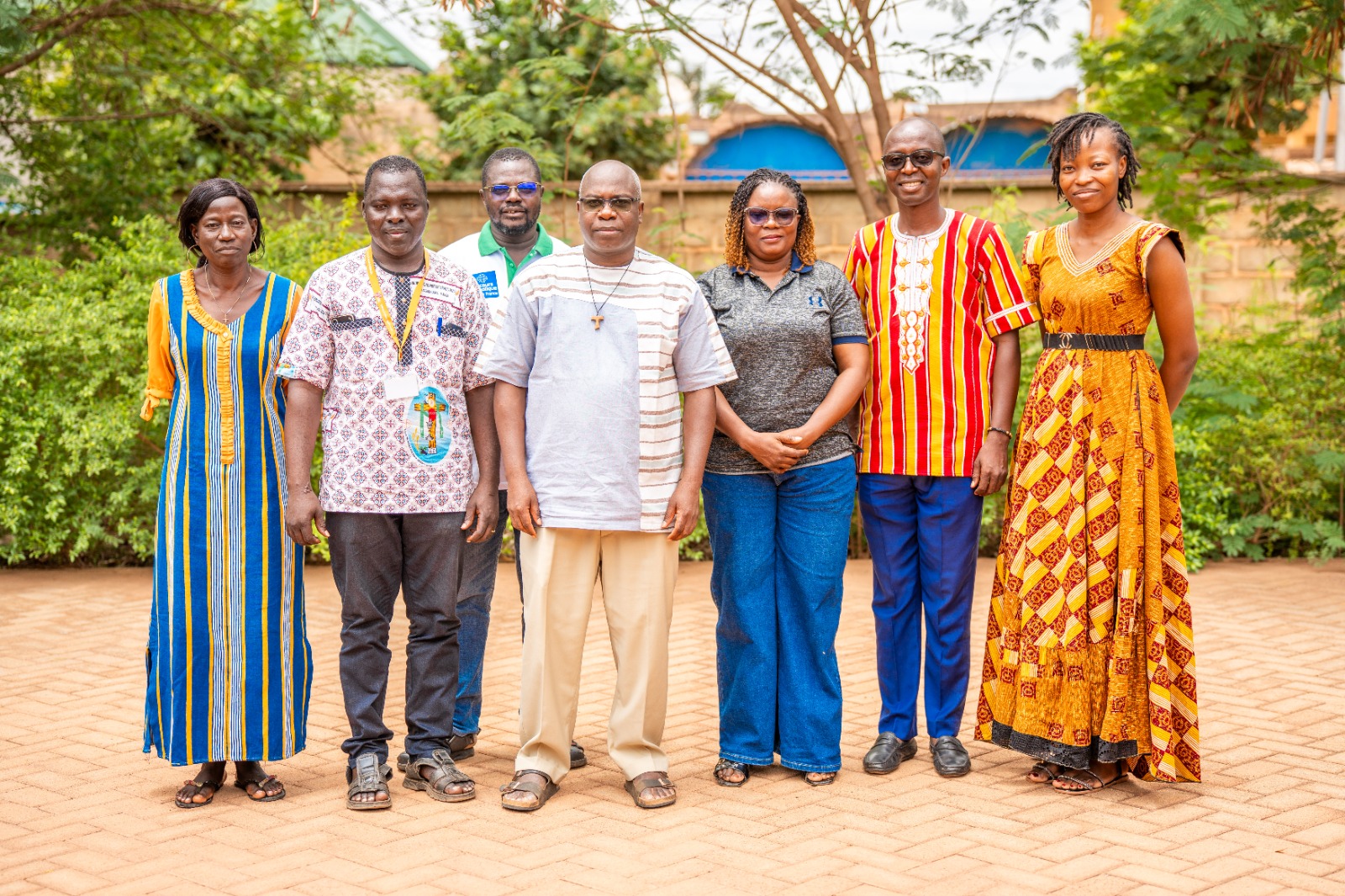 Seven adults stand in a row outdoors, wearing bright, colorful traditional attire in a garden setting.
