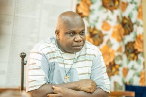 WhatsApp Image 2026-07-17 at 193703 - Tous pour l'homme, chacun pour le faible Man with a shaved head in a light blue striped shirt, seated at a table and looking to the side, wearing a cross necklace.