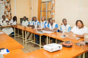 Group of diverse adults seated around a long wooden conference table in a classroom, listening attentively during a meeting, some with laptops and notebooks in front of them.