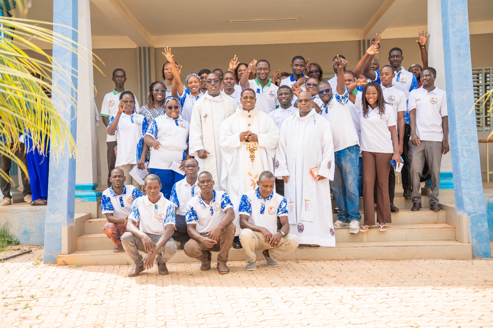 Group photo of a large community on building steps, with two priests in white robes at center and members in white/blue shirts waving to the camera.