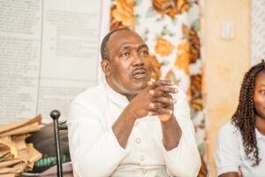 Man in a white traditional shirt sits with clasped hands and a thoughtful expression, floral wallpaper in the background.