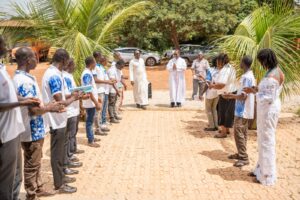People form two facing lines as a priest leads an outdoor ceremony under palm trees on a brick path.