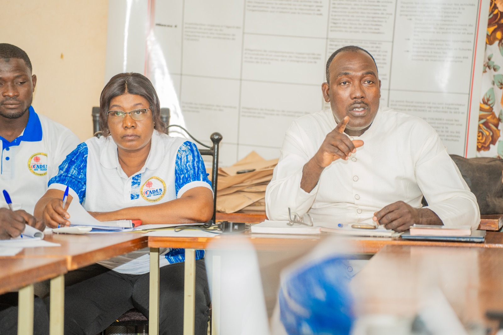 Group of people seated at a meeting table; woman in the center wears glasses and a blue-and-white polo with a badge, one man on the right speaks and points.